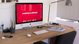 A modern workspace featuring a desktop computer with a bold red wallpaper displaying the phrase 'creativity is a lifestyle'. The desk is organized with a potted plant, books, a keyboard, and a mouse. Nearby, a sleek desk lamp provides additional lighting, and personal items like glasses and a smartphone are neatly placed.