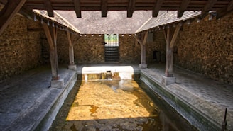 An old stone washhouse with a tiled roof structure and wooden beams, featuring a rectangular stone basin filled with water. The washhouse is surrounded by stone walls and has a cobblestone walkway along each side of the basin.