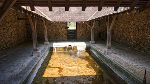 An old stone washhouse with a tiled roof structure and wooden beams, featuring a rectangular stone basin filled with water. The washhouse is surrounded by stone walls and has a cobblestone walkway along each side of the basin.