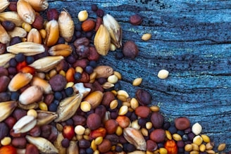 A variety of seeds and grains are scattered on a textured blue wooden surface. The seeds are diverse in size and color, ranging from small black and round to larger, oblong, and tan-colored ones. The wooden background contrasts with its rich blue tones and visible grains.