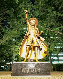 A bronze statue of a character stands prominently, striking a triumphant pose with one arm raised. The character wears a hat, an open shirt, shorts, and a flowing cape. It is set against the backdrop of lush green trees and some building lights visible through the foliage.