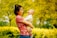 A woman in a red patterned shirt is holding a laughing baby outdoors surrounded by bright yellow and green foliage.