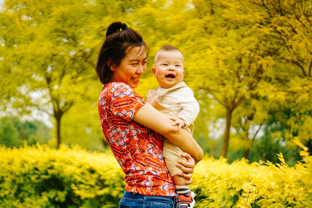 woman holding smiling baby near trees
