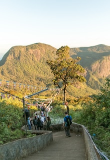 A stone pathway leads downhill, bordered by greenery and a few trees. Colorful prayer flags hang across the path. Several people walk leisurely along the pathway, enjoying a scenic view of verdant mountains in the background, illuminated by soft sunlight.