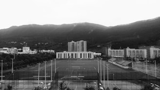 An open sports field is surrounded by several multi-story buildings, likely part of a school or university campus. The field includes tracks and is equipped with goalposts. In the background, a range of forested hills creates a natural backdrop beneath a cloudy sky.