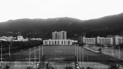 An open sports field is surrounded by several multi-story buildings, likely part of a school or university campus. The field includes tracks and is equipped with goalposts. In the background, a range of forested hills creates a natural backdrop beneath a cloudy sky.
