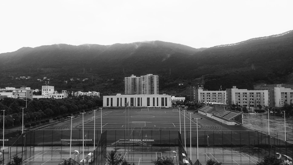 An open sports field is surrounded by several multi-story buildings, likely part of a school or university campus. The field includes tracks and is equipped with goalposts. In the background, a range of forested hills creates a natural backdrop beneath a cloudy sky.