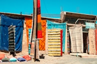 Colorful handmade rugs and carpets hang on a wall in an outdoor market setting. The assortment includes various vibrant hues and patterns. Pillows are laid out on the ground, adding to the display of textiles. The backdrop is a rustic wall with a small sign visible amongst the fabrics.