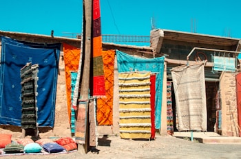 Colorful handmade rugs and carpets hang on a wall in an outdoor market setting. The assortment includes various vibrant hues and patterns. Pillows are laid out on the ground, adding to the display of textiles. The backdrop is a rustic wall with a small sign visible amongst the fabrics.