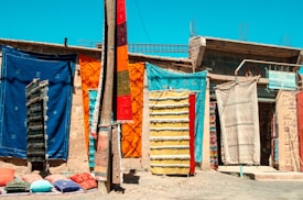 Colorful handmade rugs and carpets hang on a wall in an outdoor market setting. The assortment includes various vibrant hues and patterns. Pillows are laid out on the ground, adding to the display of textiles. The backdrop is a rustic wall with a small sign visible amongst the fabrics.