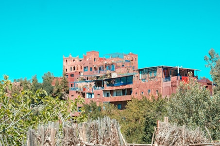 A cluster of traditional mud-brick houses is situated on a hillside surrounded by lush greenery. The structures exhibit a range of earthy colors and have flat rooftops, with some featuring wooden or thatch shades. The bright, clear sky provides a vivid blue backdrop.