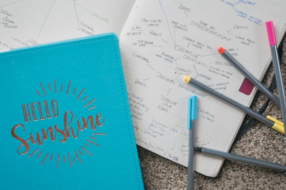 A blue notebook with 'Hello Sunshine' written on its cover in elegant lettering sits beside an open planner filled with handwritten notes and diagrams. Several colored pens, including yellow, orange, blue, and pink, are scattered across the pages and carpet.