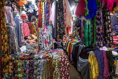 A colorful fabric store filled with various vibrant textiles. The fabrics are hanging and stacked all around, displaying an array of patterns and colors. Amongst the fabrics, a person is resting, lying down on a pile of materials, appearing relaxed amidst the bustling market environment.