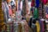 A colorful fabric store filled with various vibrant textiles. The fabrics are hanging and stacked all around, displaying an array of patterns and colors. Amongst the fabrics, a person is resting, lying down on a pile of materials, appearing relaxed amidst the bustling market environment.