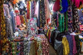 A colorful fabric store filled with various vibrant textiles. The fabrics are hanging and stacked all around, displaying an array of patterns and colors. Amongst the fabrics, a person is resting, lying down on a pile of materials, appearing relaxed amidst the bustling market environment.
