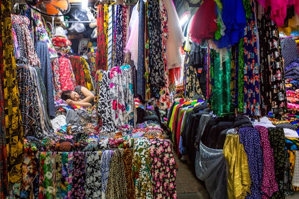 A colorful fabric store filled with various vibrant textiles. The fabrics are hanging and stacked all around, displaying an array of patterns and colors. Amongst the fabrics, a person is resting, lying down on a pile of materials, appearing relaxed amidst the bustling market environment.