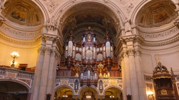 An ornate and grand architectural interior featuring an elaborate pipe organ as the focal point. The surrounding walls and ceiling are richly decorated with intricate carvings and lavish details. Soft lighting highlights the classical columns and decorative arches.