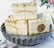 Several bars of handmade soap are neatly stacked on a scalloped metal dish. The soaps are individually wrapped in clear packaging and tied with rustic twine. A round label on one of the soaps reads 'Hand Crafted with Love For You.' The background features floral elements on a light surface.