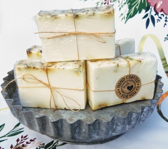 Several bars of handmade soap are neatly stacked on a scalloped metal dish. The soaps are individually wrapped in clear packaging and tied with rustic twine. A round label on one of the soaps reads 'Hand Crafted with Love For You.' The background features floral elements on a light surface.