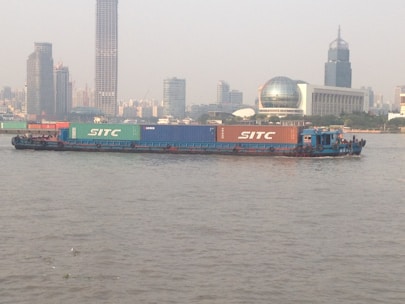 A large barge carrying multiple shipping containers is seen floating on a river against a backdrop of a city skyline. The containers have the word 'SITC' written on them. Various high-rise buildings, including a tower and a dome-like structure, are visible in the background under a hazy sky.