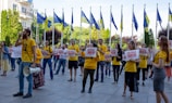 A group of people wearing yellow shirts are participating in a protest or demonstration. They are holding signs that include the number 10019 and are gathered in an outdoor setting. European Union and national flags are visible in the background, alongside trees and buildings. A man is speaking into a megaphone.