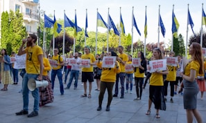 A group of people wearing yellow shirts are participating in a protest or demonstration. They are holding signs that include the number 10019 and are gathered in an outdoor setting. European Union and national flags are visible in the background, alongside trees and buildings. A man is speaking into a megaphone.