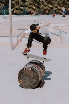 A skateboarder performing an aerial trick over a graffiti-covered barrel in an outdoor skate park. The individual is dressed in casual attire, including a white shirt, black pants, and red sneakers, and is mid-air, capturing a moment of action and skill. The background shows palm trees and other people in the skate area.