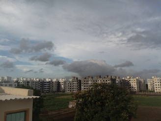 A residential area with modern apartment buildings is seen under a cloudy sky. In the foreground, there is a large tree and patches of green fields. The skyline is dominated by grey and white clouds, creating a contrast with the structures.