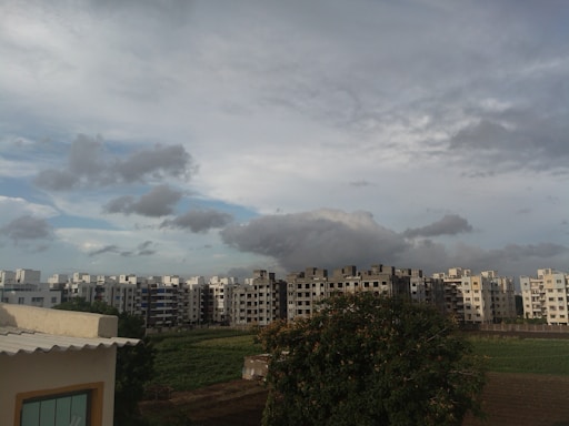 A residential area with modern apartment buildings is seen under a cloudy sky. In the foreground, there is a large tree and patches of green fields. The skyline is dominated by grey and white clouds, creating a contrast with the structures.