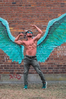 A shirtless man poses confidently in front of a brick wall adorned with graffiti art depicting large, vibrant green wings. He wears sunglasses, jeans, and teal sneakers, flexing his muscles dramatically as if the wings belong to him.