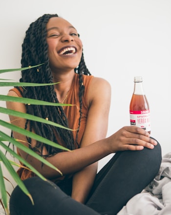 A person with braided hair is laughing joyfully while holding a bottle of sparkling yerba mate beverage. They are seated with one knee bent and wearing a sleeveless brown top. A green plant is partially visible in the foreground.