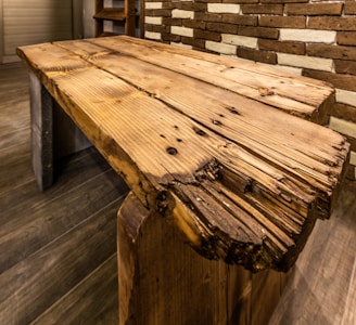 A rustic wooden table made of thick, rough-hewn planks with visible knots and imperfections. The table is set against a background with a textured brick wall, some bricks painted white, and a wooden floor. The table's edges are uneven and worn, adding to its antique appearance.