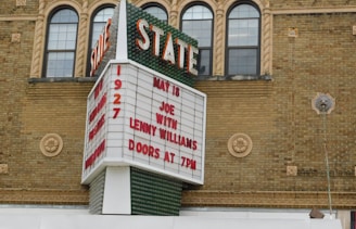 A historic theater marquee with classic architectural elements and a sign advertising an event featuring Joe and Lenny Williams on May 18, with doors opening at 7 PM. The marquee displays large, bold red lettering against a white background, set against a detailed brick facade with arched windows and decorative stonework.