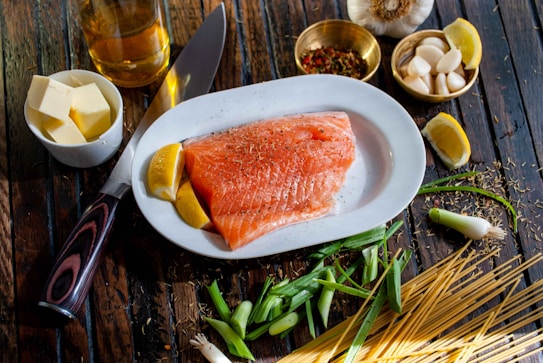 A raw salmon fillet seasoned with herbs is placed on a white plate with lemon wedges. Surrounding the plate are various cooking ingredients including a knife, butter cubes in a bowl, a bottle of oil, garlic cloves, colorful spices in a brass bowl, chopped green onions, and uncooked spaghetti noodles scattered on a rustic wooden table.
