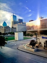 A modern urban cityscape featuring tall buildings with glass facades, with a prominent triangular blue building. The sunset casts a warm reflection on one of the glass buildings, adding a glow to the scene. In the foreground, there's an outdoor seating area next to a building labeled 'Biergarten'. Trees line the streets, and a rooftop-like green area is visible nearby.