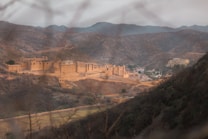 A large, historic fort with intricate architecture is nestled among rugged hills. The fort's sandy color blends with the landscape, and pathways are visible leading to and surrounding the structure.