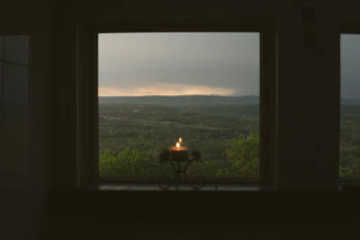A cozy candle glowing softly on a wooden table with a backdrop of rolling Kentucky bluegrass hills at sunset.