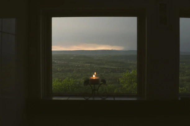 A cozy candle glowing softly on a wooden table with a backdrop of rolling Kentucky bluegrass hills at sunset.