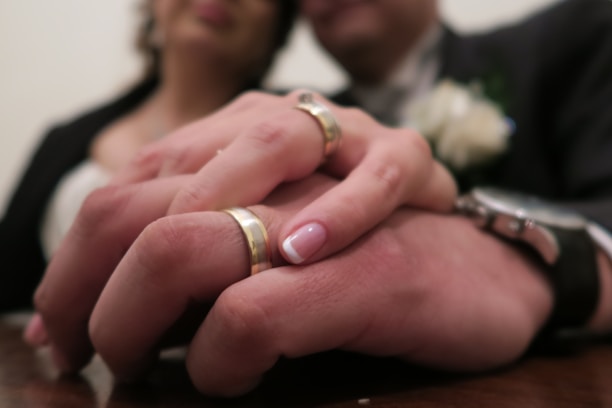 A close-up of two hands wearing wedding rings, gently clasping each other, with a blurred background of a couple. The woman's manicured nails are visible, and there is a soft focus on a bouquet near the man's lapel.