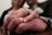 A close-up of two hands wearing wedding rings, gently clasping each other, with a blurred background of a couple. The woman's manicured nails are visible, and there is a soft focus on a bouquet near the man's lapel.
