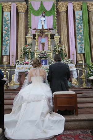 A bride and groom kneel at an ornate altar adorned with floral arrangements and religious icons. The bride is wearing a white gown with a long train and a veil, while the groom is dressed in a dark suit. The altar is richly decorated with gold accents, colorful drapery in green and pink, and religious statues including a crucifix.