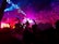 A crowded concert or club scene with vibrant colors and energetic lighting. People are enthusiastically raising their hands amidst a shower of confetti. The stage is illuminated with bright lights, creating a dynamic and festive atmosphere.