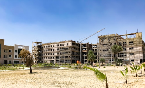 A construction site with several multi-story buildings under development. Scaffolding is visible around the structures, and a large crane hovers above. Palm trees are planted in the sandy foreground, with the clear blue sky providing a backdrop.