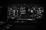 A dimly lit retail display features various merchandise neatly arranged on shelves, with a prominent neon sign reading 'OUT, DAMNED SPOT!' The shelves are filled with books, trinkets, and other small items, casting a moody, atmospheric vibe.
