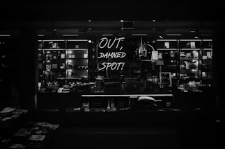 A dimly lit retail display features various merchandise neatly arranged on shelves, with a prominent neon sign reading 'OUT, DAMNED SPOT!' The shelves are filled with books, trinkets, and other small items, casting a moody, atmospheric vibe.