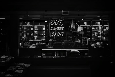 A dimly lit retail display features various merchandise neatly arranged on shelves, with a prominent neon sign reading 'OUT, DAMNED SPOT!' The shelves are filled with books, trinkets, and other small items, casting a moody, atmospheric vibe.