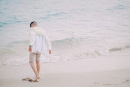 A young child walks barefoot along a sandy beach, near the gentle waves of the sea. The child's attire includes a light jacket and rolled-up pants, suggesting a casual and relaxed atmosphere.