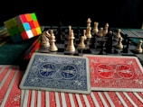 A colorful Rubik's cube sits next to a chessboard with pieces arranged for a game. In the foreground, there are several playing cards with intricate red and blue designs.