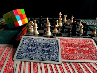 A colorful Rubik's cube sits next to a chessboard with pieces arranged for a game. In the foreground, there are several playing cards with intricate red and blue designs.