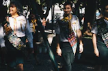 A group of people wearing uniforms adorned with numerous badges, indicating some form of achievement or participation, possibly related to scouting or similar organizations. The scene is set outdoors in a sunlit park area with trees providing shade. The individuals appear to be walking in a relaxed and friendly manner.